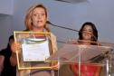 Aparecida Benito Pereira pronunciando, emocionada, sobre o prêmio recebido.
Ao Fundo Mira Graçano, reporter e apresentadora da RPC TV, afiliada da Rede Globo.