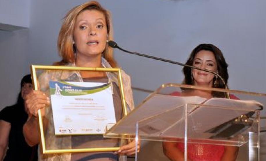 Aparecida Benito Pereira pronunciando, emocionada, sobre o prêmio recebido.
Ao Fundo Mira Graçano, reporter e apresentadora da RPC TV, afiliada da Rede Globo.